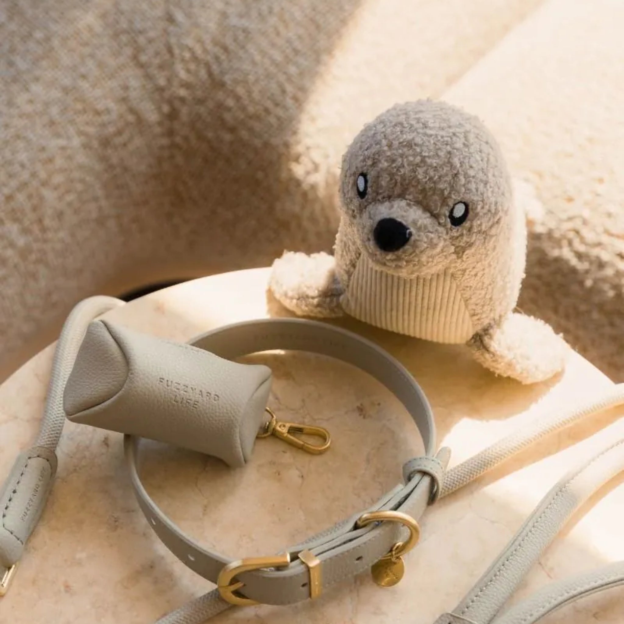 A warm lifestyle shot of the FuzzYard Life Sandstone Seal plush dog toy sitting on a beige marble table next to a light grey leather dog collar and a matching leather poop bag dispenser pouch.