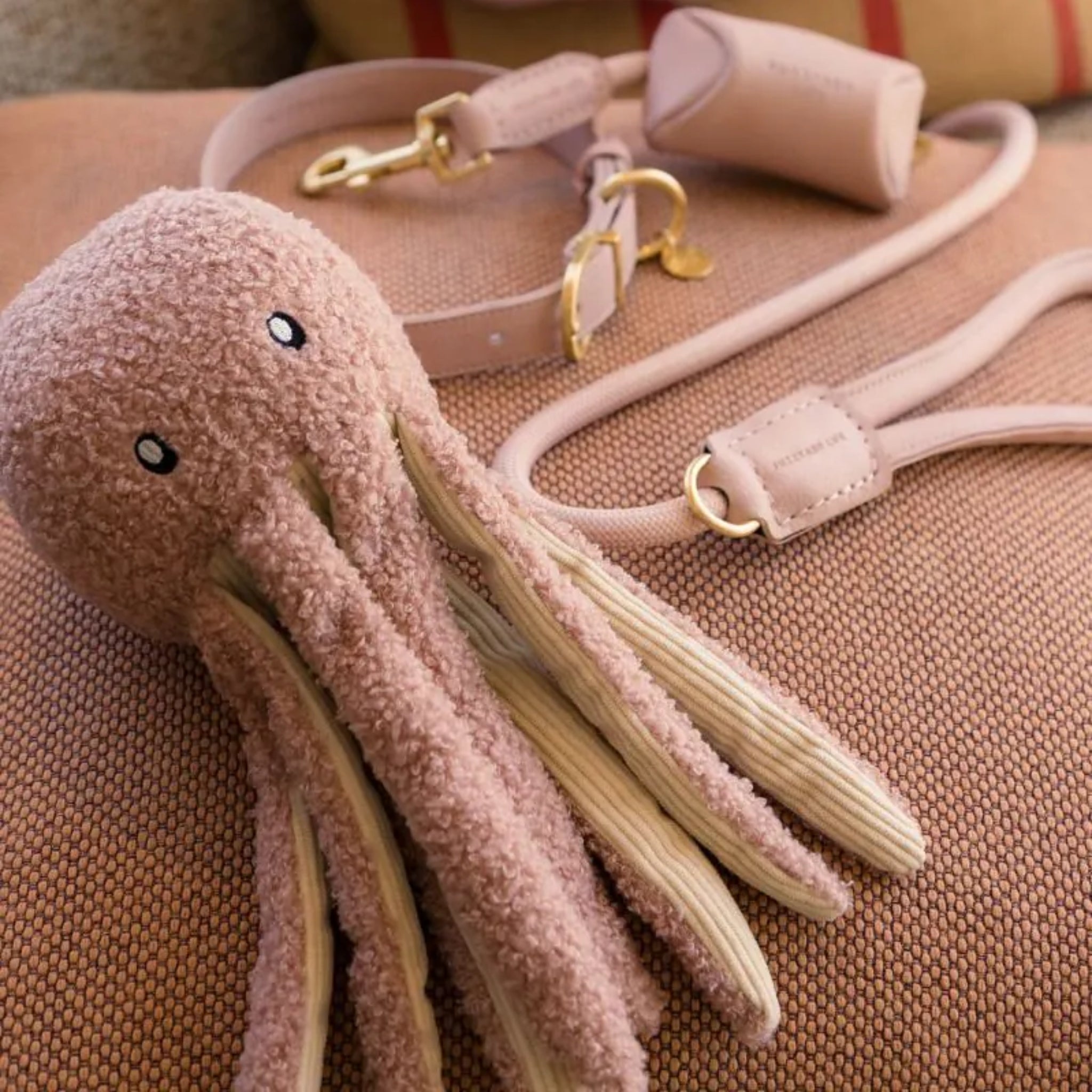 A close-up lifestyle shot of the FuzzYard Life Soft Blush Octopus dog toy on a textured cushion, showing its embroidered eyes and cream corduroy tentacle undersides next to a matching blush pink leather collar and leash set.