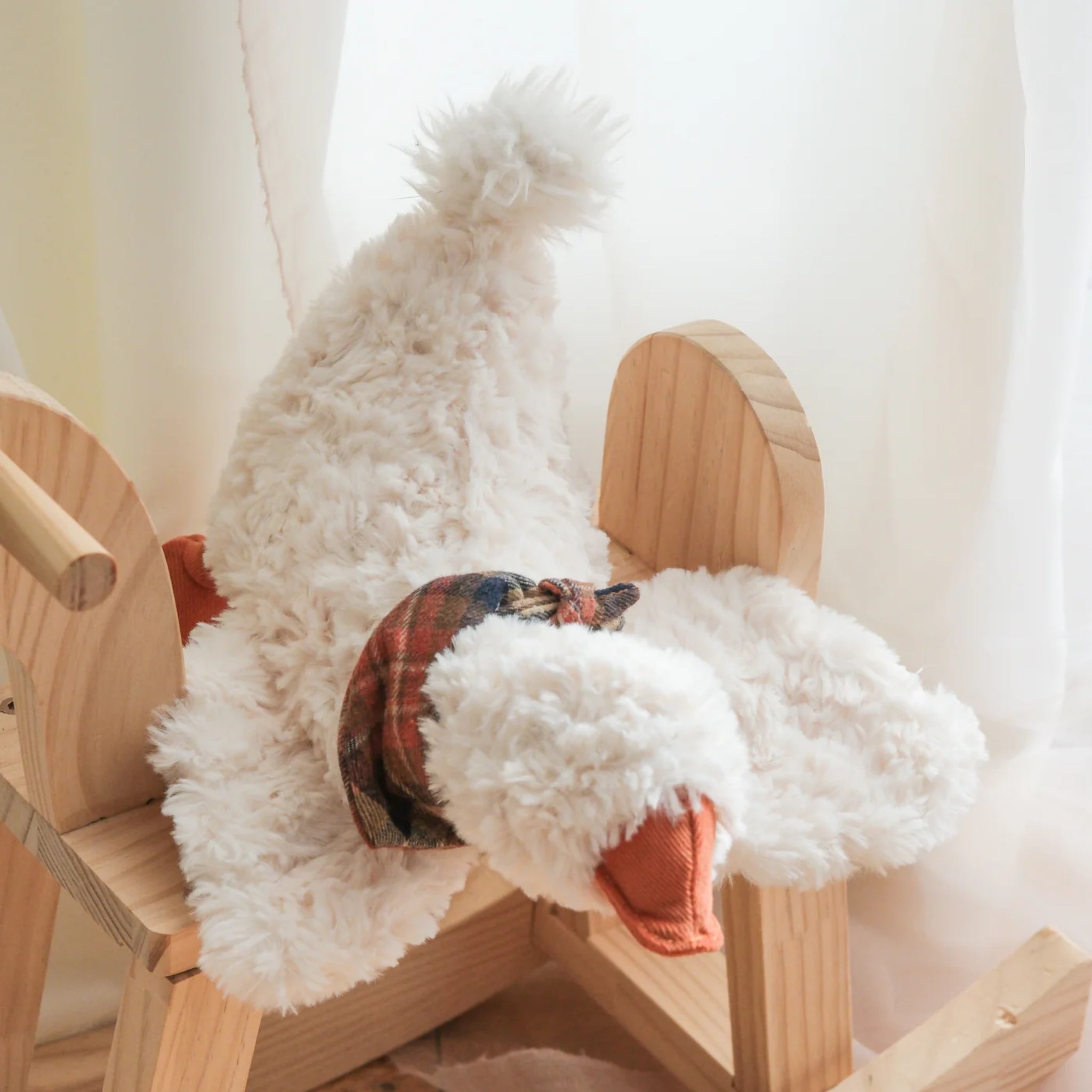 The textured white plush goose dog toy draped over a small wooden rocking horse indoors against a bright, soft-lit window backdrop.