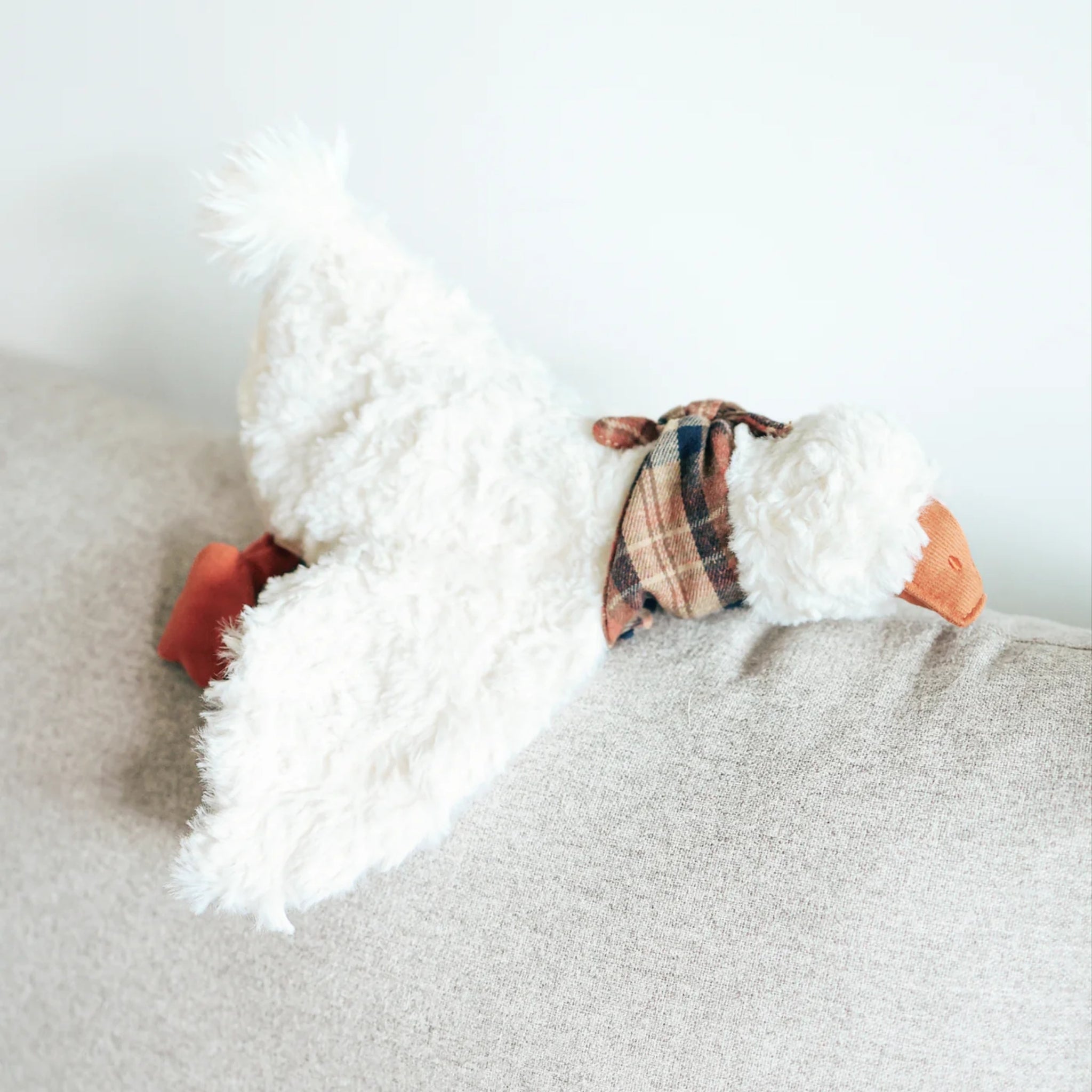 A close-up view of the Gus the Goose plush pet toy lying across the back of a grey sofa, highlighting its textured white faux-fur body, orange corduroy beak, and brown plaid scarf.