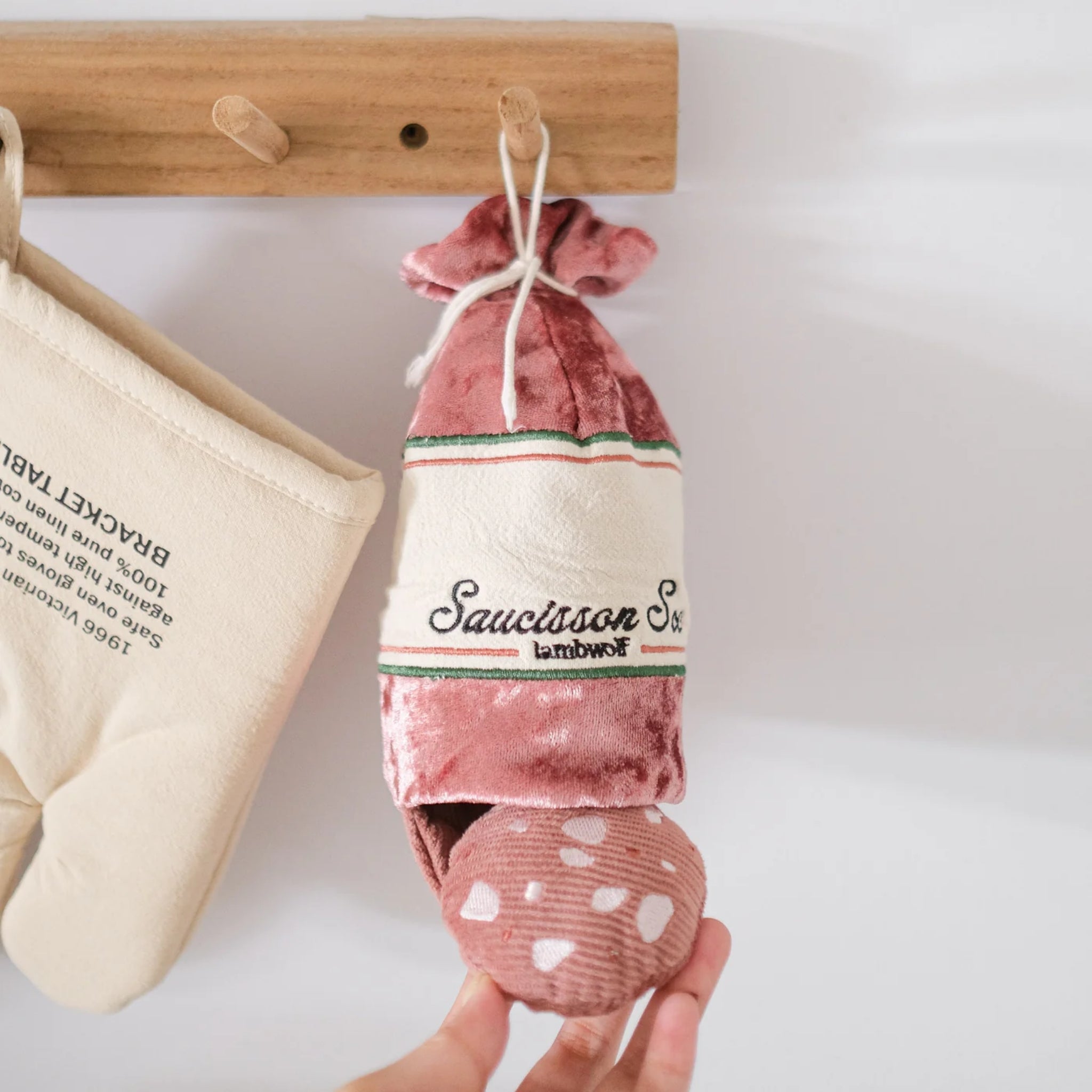 Close-up of a hand gently reaching for the Lambwolf Collective Saucisson Sec plush toy, which is hanging from a wooden wall peg using its white top string alongside a canvas oven mitt.