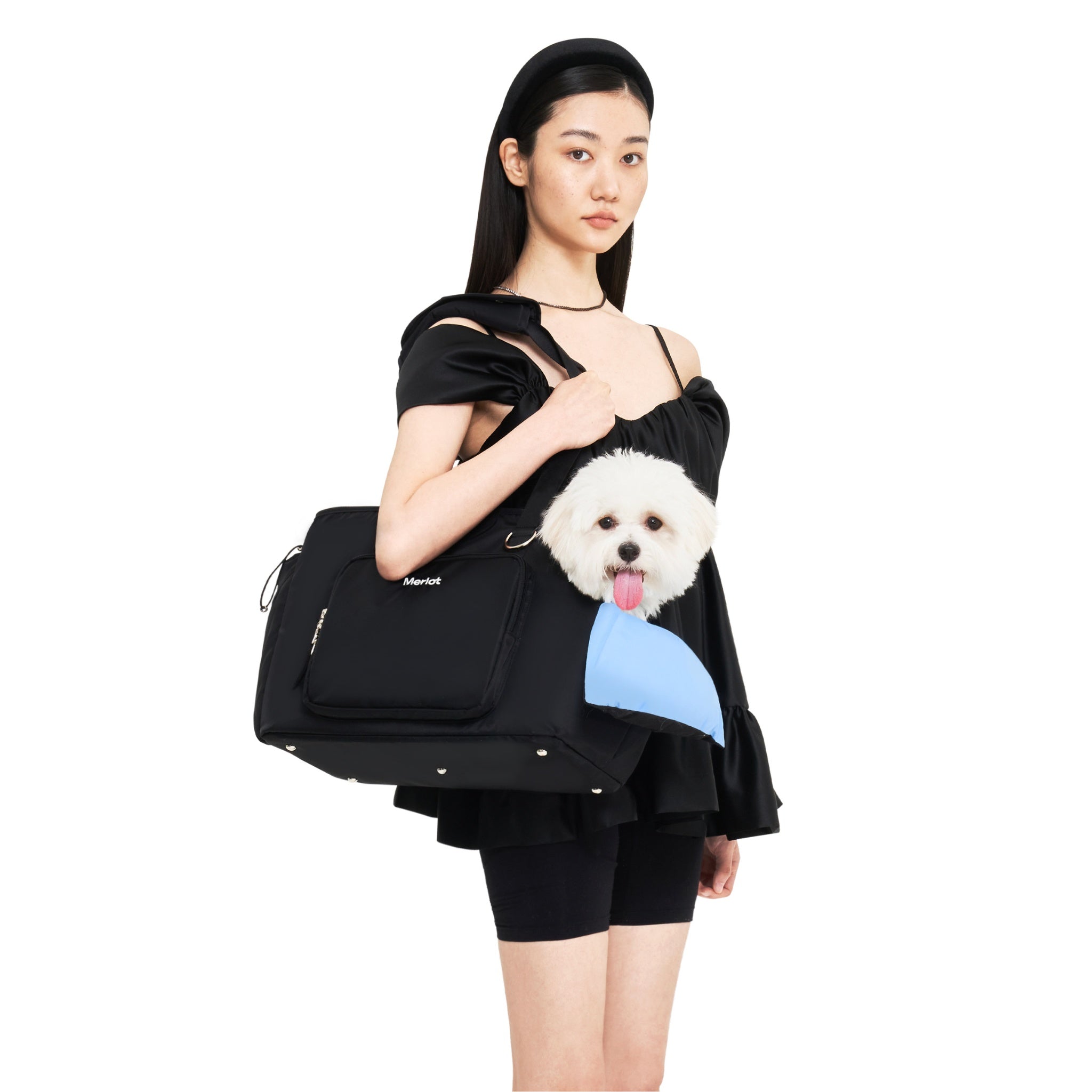 A model in a black dress carrying the black Merlot Mermer dog carrier bag on her shoulder, with a small white Maltese dog looking out over a light blue comfort cushion.