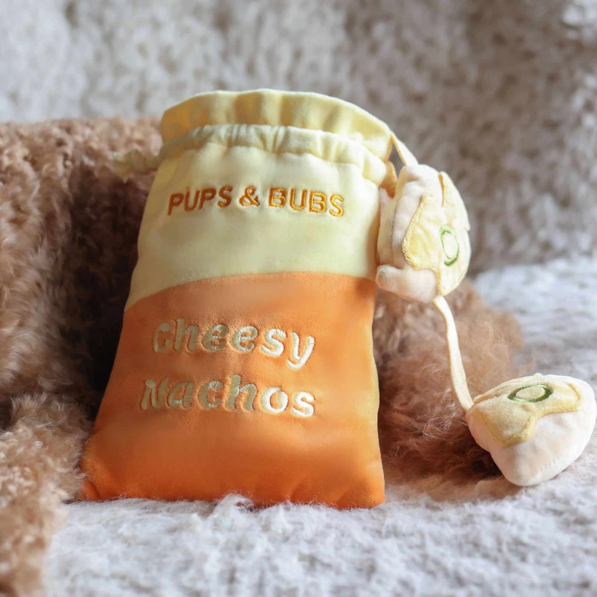 Close-up shot of the yellow and orange Pups and Bubs Cheesy Nachos plush toy bag resting on a soft, textured white blanket with a poodle in the background.