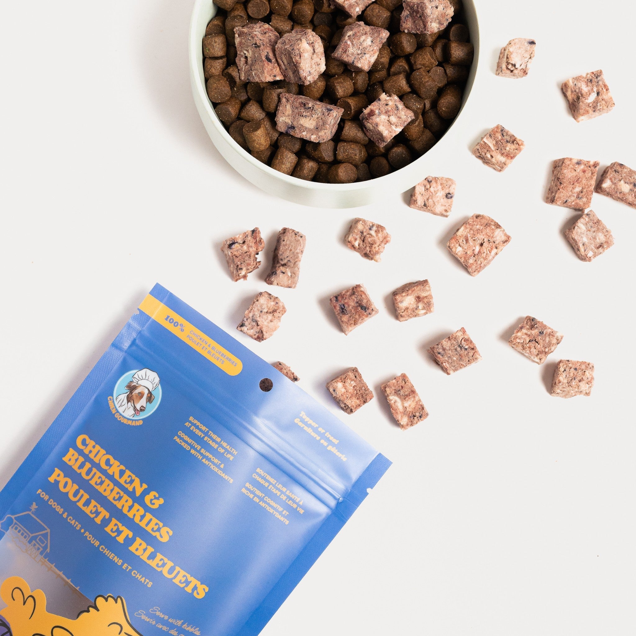 A top-down view of Chien Gourmand freeze-dried chicken and blueberry cubes being used as a pet food topper, scattered around and inside a food bowl filled with brown kibble next to its blue packaging.