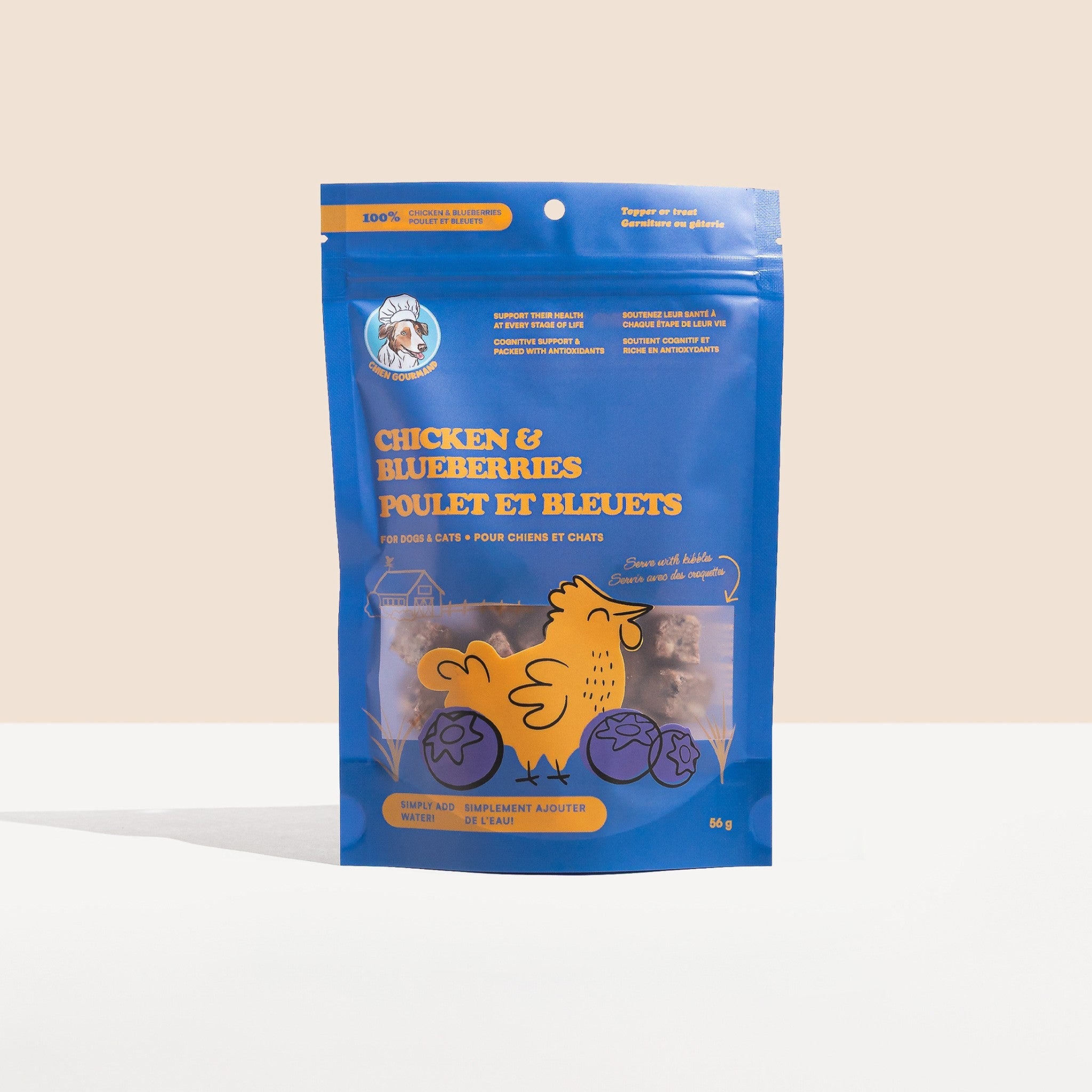 A studio shot of Chien Gourmand's blue stand-up pouch for Chicken and Blueberries freeze-dried dog and cat treats, featuring a bright yellow chicken and blueberry graphic over a clear window showing the food cubes.