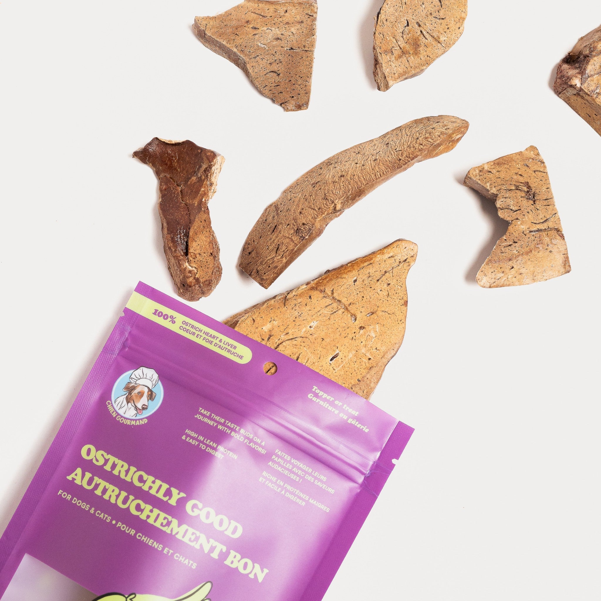 A top-down lifestyle view showing freeze-dried ostrich heart and liver pieces tumbling out from a purple Chien Gourmand product pouch onto a clean white surface.