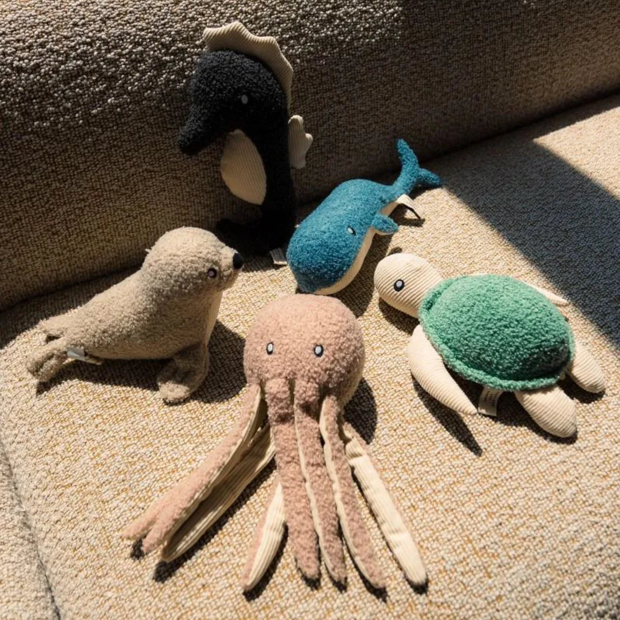 A group lifestyle shot of the FuzzYard Life ocean plush dog toy collection resting on a textured beige fabric sofa, including the sandstone seal, slate grey seahorse, French blue whale, blush octopus, and myrtle green turtle.