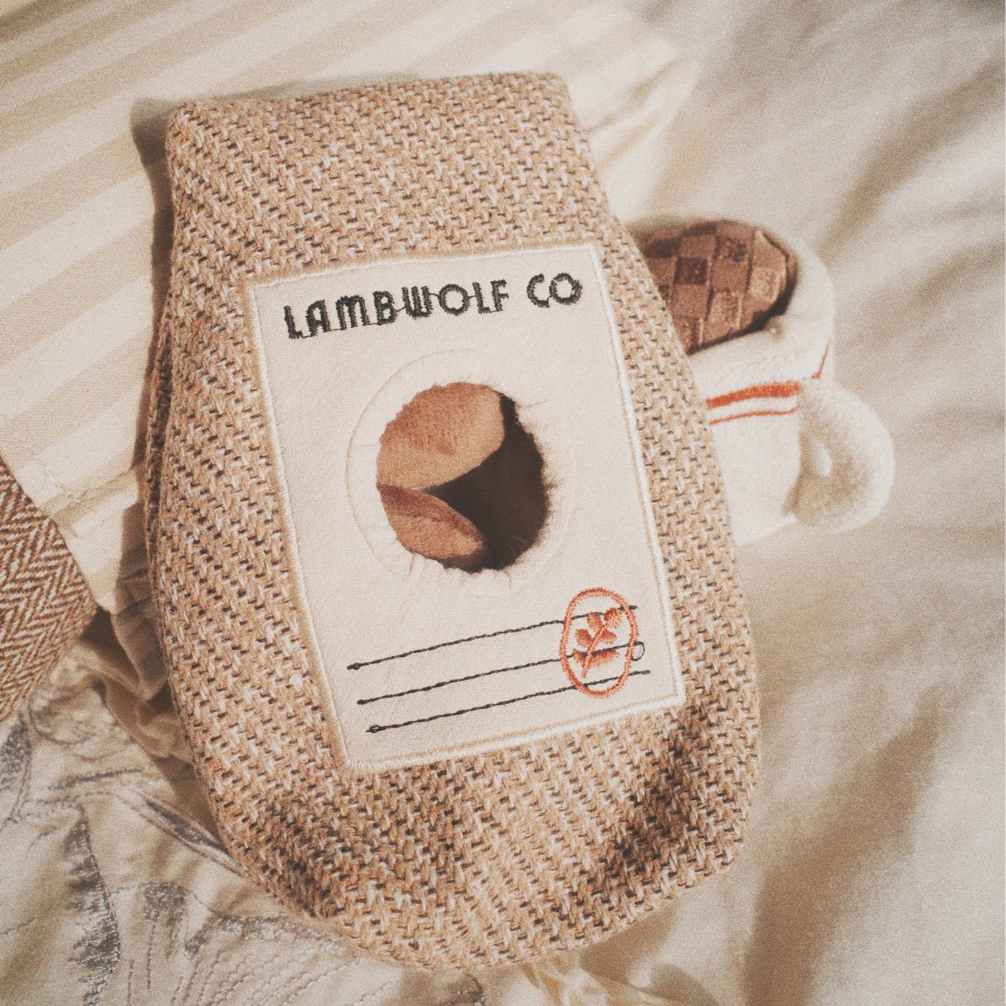 A close-up view of the textured woven fabric coffee bag plush toy with a circular front opening showing brown plush coffee beans nestled inside.