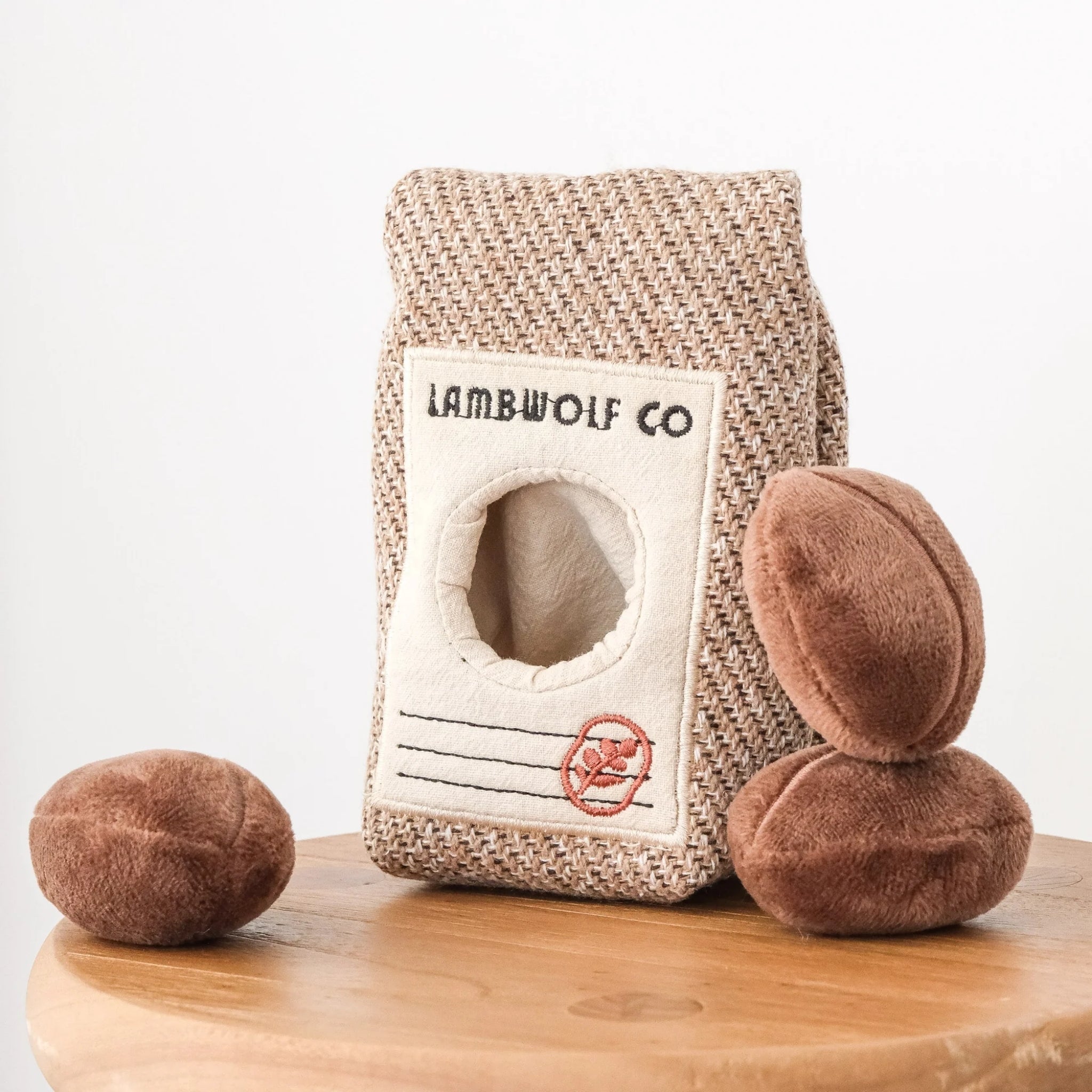 The Lambwolf Co woven coffee bean bag puzzle toy standing on a round wooden stool alongside three separate plush brown squeaky coffee beans.