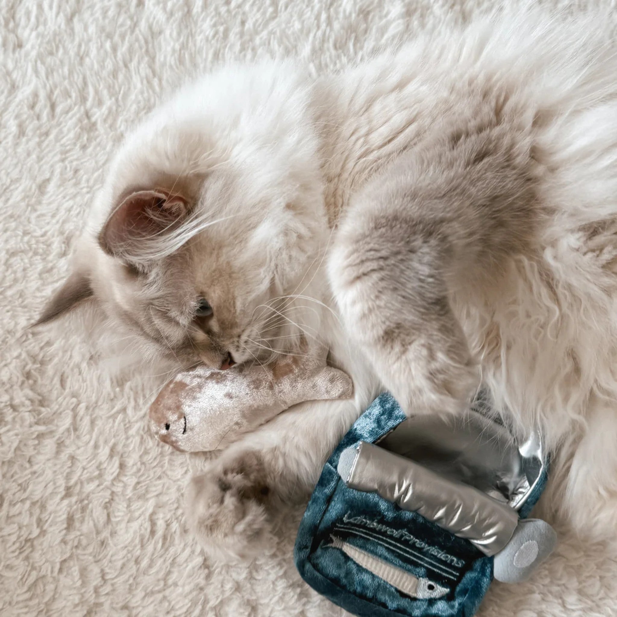 A long-haired cream cat rolling on its back on a fuzzy rug, cuddling and licking a small velvet plush sardine fish toy filled with catnip.