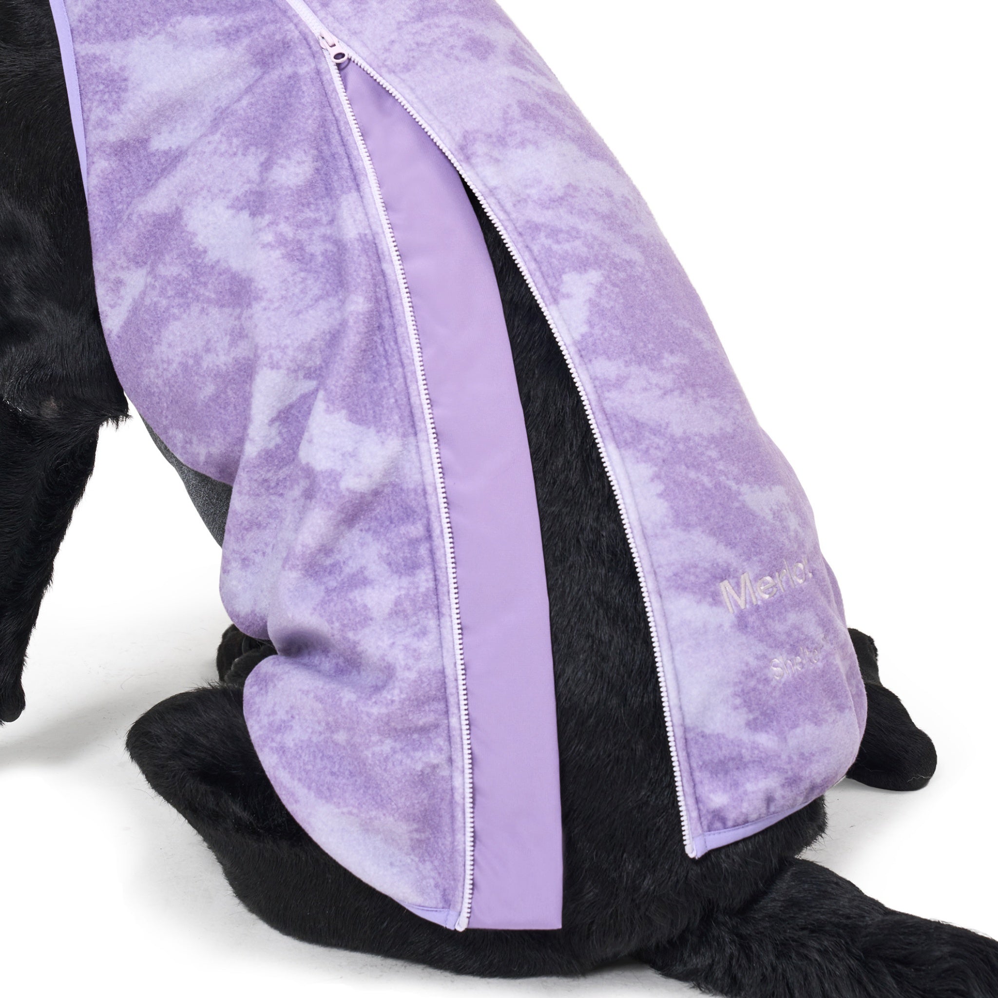 Rear view of a large black Labrador sitting down, showcasing the functional back zipper with an integrated protective fabric guard and embroidered brand logo on the purple tie-dye fleece Merlot Shelter jacket.