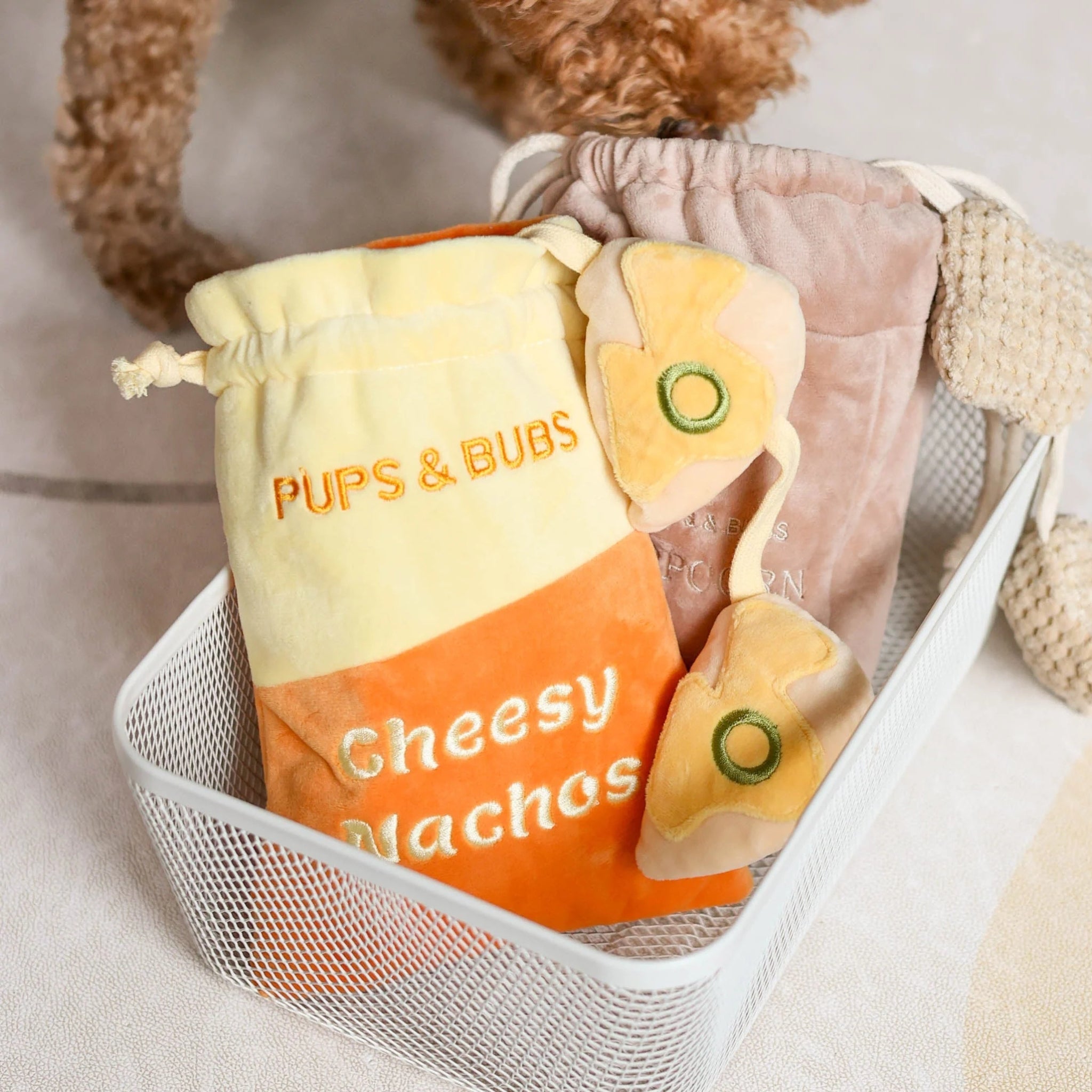 The Pups and Bubs Cheesy Nachos interactive plush dog toy bag sitting in a white wire basket next to a plush popcorn bag toy.