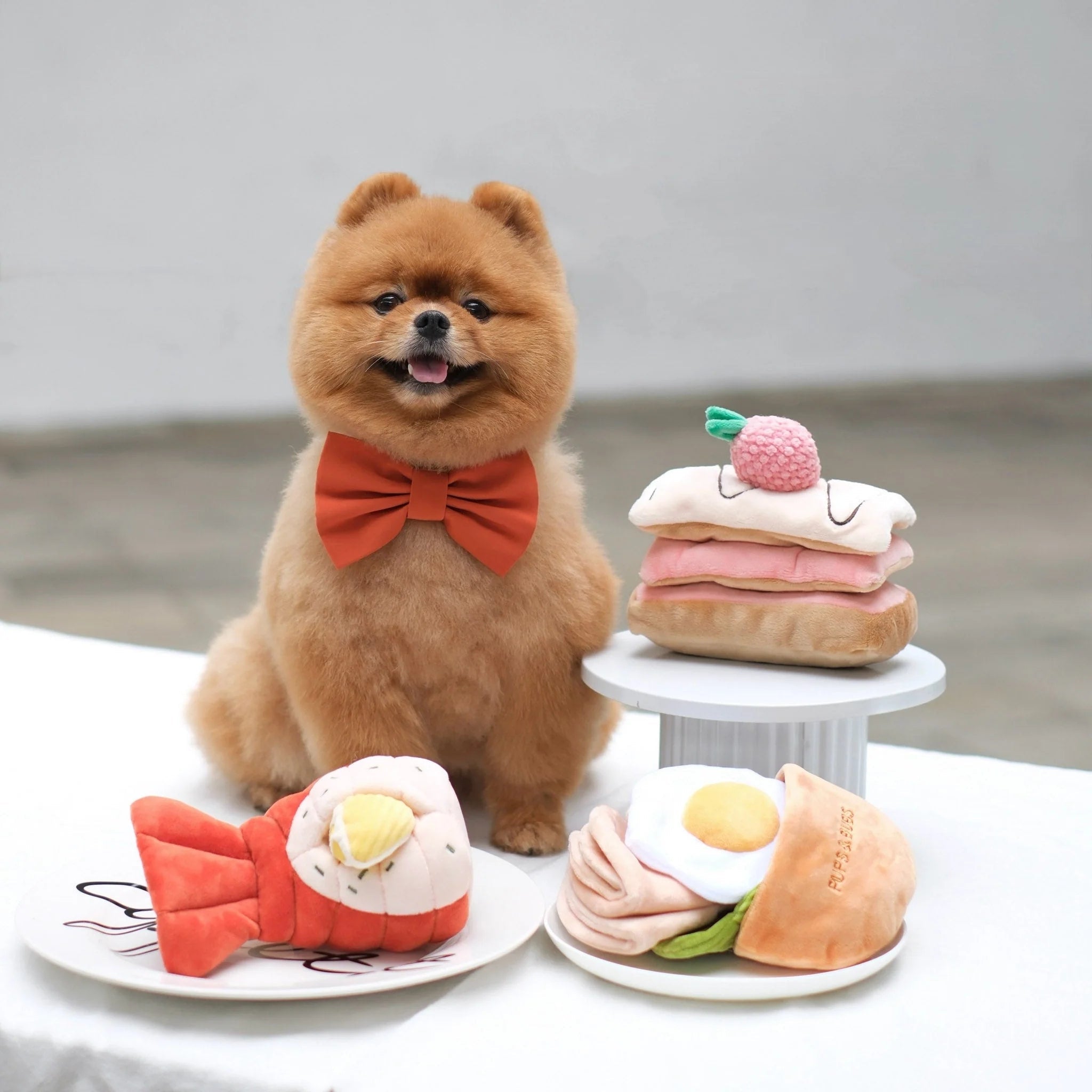 A Pomeranian dog wearing an orange bow tie sitting behind a collection of Pups and Bubs plush interactive toys, including the grilled lobster, an egg pita pocket, and a cake slice.